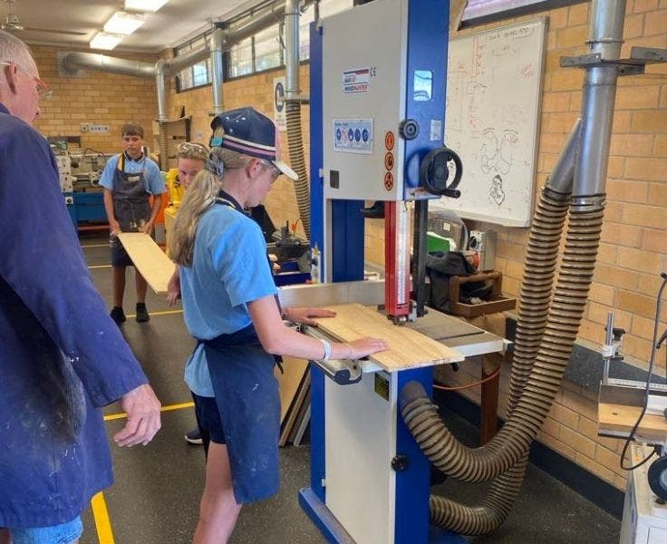 A student using a wood bandsaw in the construction workshop under the careful supervision of the woodwork teacher