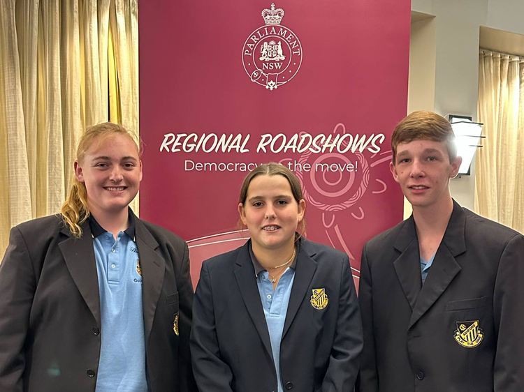 Three students wearing blazers standing in front of the NSW Parliament regional roadshow democracy in action signgn