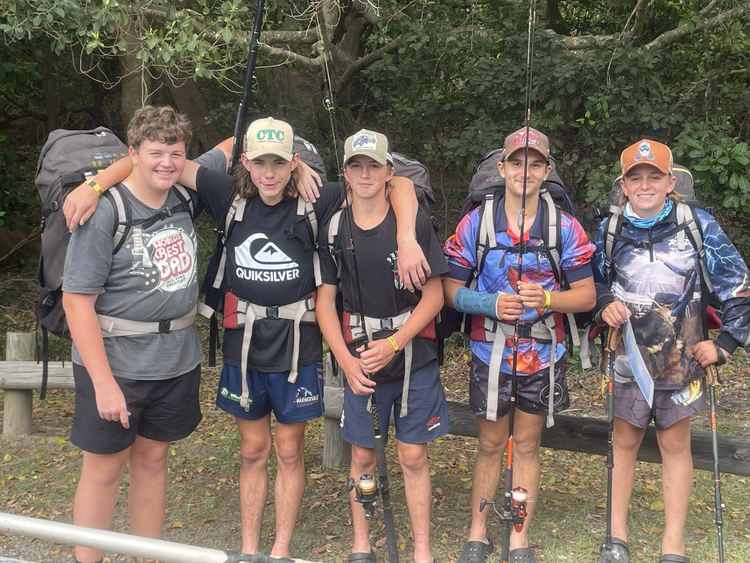 Five students who are wearing shorts and fishing shirts standing in a line holding fishing rods
